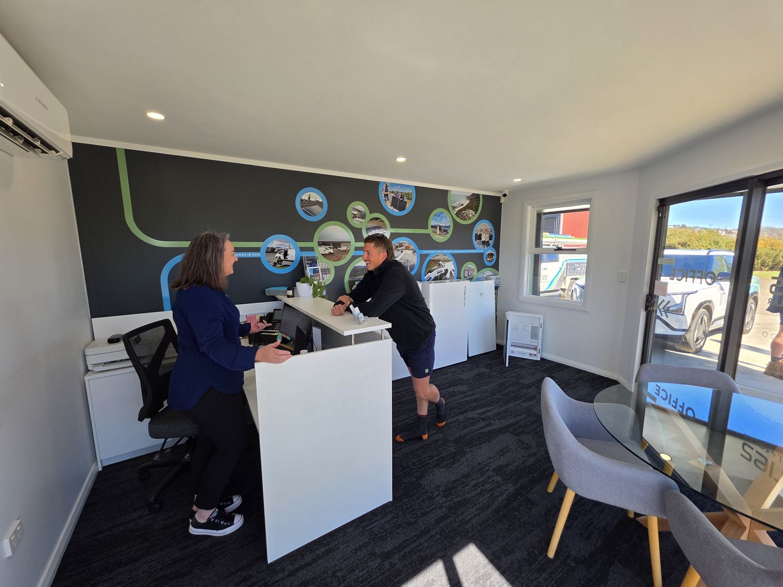 Kel and Brad at the In Series office reception desk, with the office's Tasmania-themed circle mural on the wall behind them